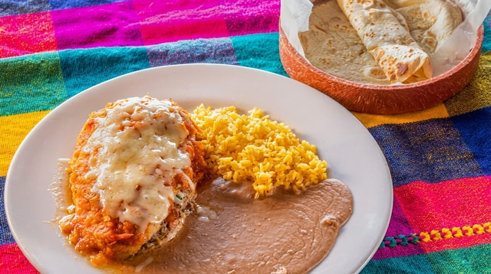 A plate of Mexican food with a chile relleno, yellow rice, refried beans, and a basket of tortillas.